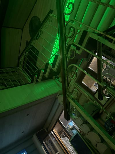 An upward-angled interior photograph depicts a multi-level space illuminated by a prominent green LED light strip. A wooden staircase with ornate metallic railings featuring scrollwork ascends diagonally from the lower left towards the upper right. The green light casts a strong luminescence across the wooden planked walls and ceiling. The underside of the stairs exhibits a distinct zigzag architectural pattern. In the upper left background, a dark circular object is mounted on a wooden wall. Windows with dark wooden frames are visible through and behind the staircase on the right. In the lower right foreground, decorative wooden log slices and a dark rectangular panel are present. Further to the left and below, an additional window and a white wall-mounted appliance are partially visible. No individuals or active interactions are depicted within the frame. This scene is located within Matveevskoe, Russia.