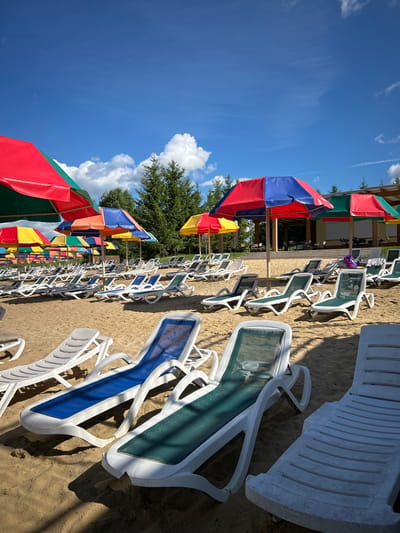 The image depicts a sandy beach with numerous empty lounge chairs arranged in rows under colorful umbrellas. The umbrellas are a mix of red and blue, red and green, and yellow and red, providing shade on a sunny day. The beach is backed by a line of trees and a building with a covered outdoor seating area. The sky is clear blue with a few scattered white clouds. The overall impression is one of a recreational area, likely a public beach or resort, on a pleasant day. It is not possible to determine the specific location within Yubileynyy, Russia, based on the visual information provided.