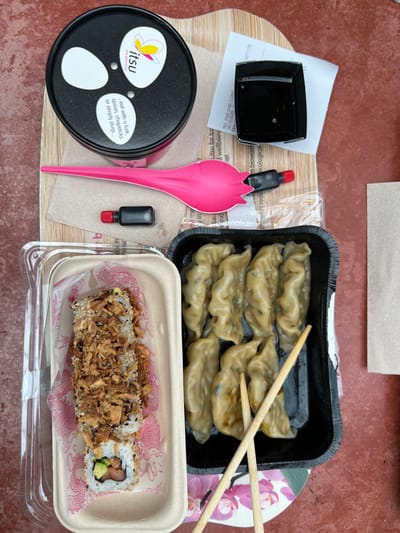 An overhead photograph depicts a pre-packaged meal from "itsu" laid out on a reddish-brown textured surface, presumably in Steenokkerzeel, Belgium. The primary subjects are various food items and their accompanying utensils and condiments, arranged on a light-colored wooden board or tray.

On the left, a rectangular, light-brown biodegradable container with a clear plastic lid holds a sushi roll. The roll, cut into segments, rests on pink paper patterned with a dragon motif and is heavily topped with crispy fried onions and white sesame seeds. Visible at the end are rice, nori, avocado, and a dark pink filling.

In the upper central area, a black cylindrical cup with a black lid features the "itsu" logo and butterfly graphic, alongside text reading, "eat with a fork, spoon, chopsticks or simply slurp...". Below it, on a light-brown paper napkin, lies a bright pink spork-like utensil with a black handle tip and red stopper, accompanied by a small black bottle with a red cap, likely containing soy sauce.

To the upper right, a small black square dish, containing a dark liquid, rests partially on a crumpled white paper receipt fragment that displays a date of "19/10/2023". Below this, a black rectangular plastic tray contains approximately ten to twelve light-brown dumplings. Two light-colored wooden chopsticks are placed diagonally across the dumplings. A piece of paper with a pink and green floral orchid pattern is visible beneath parts of the dumpling tray and chopsticks. A folded light-brown paper napkin is positioned to the far right. No people are visible in the static scene.