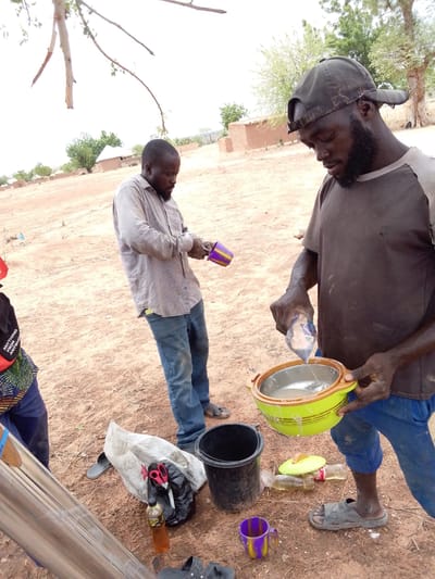 The image depicts three people in an outdoor setting that appears to be in a rural area. The ground is reddish-brown soil. In the background, there are structures that resemble traditional houses.
The main subject is a man wearing a dark t-shirt, blue pants, and a baseball cap. He is pouring liquid from a small plastic pouch into a green and yellow bowl. Another man stands to his left, wearing a collared shirt and jeans. He is holding a purple and yellow cup.
On the ground, near the man with the bowl, there is a black bucket, several plastic bottles, a pair of sandals, a purple and yellow cup, a white plastic bag with what appears to be a pair of scissors inside, and other miscellaneous items. A third person is partially visible on the left side of the frame.
The location is identified as Mulke Barikin Sayejo, Nigeria.