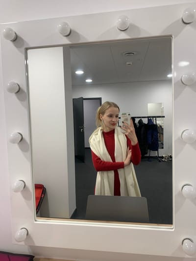 A blonde female is depicted in a mirror selfie, holding a white smartphone with dark nail polish visible on her fingers. She wears a red high-neck top and a light-colored (cream or off-white) scarf draped over her shoulders and arms, which are crossed. Her hair is pulled back in a ponytail. The reflection is captured in a large, white-framed mirror surrounded by fourteen illuminated, spherical vanity lights.
The environment reflected in the mirror appears to be a modern interior space, potentially a dressing room, located in Nowy Targ, Poland. The walls are predominantly light grey or white, and the floor is a dark grey or black carpet. To the left of the reflection, a dark grey door with a silver handle is visible. In the foreground, a dark grey chair or stool is partially obscured by the bottom frame of the mirror. To the right, a black clothing rack holds several dark-colored garments, including jackets or coats. Above the clothing rack, a smaller, frameless mirror is mounted on the wall. The ceiling features recessed lighting and a ventilation grate. A red bag or case is partially visible on the floor to the bottom left, outside the main mirror's reflection but within the overall image frame.