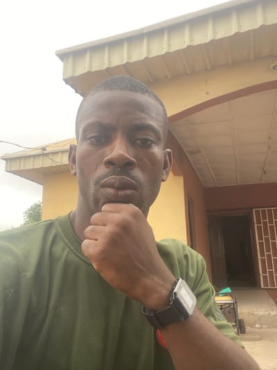 The image portrays a male individual, observed from the chest up, outdoors in Kado, Nigeria. The subject has dark skin, short dark hair, and a mustache and goatee. He looks directly at the camera with a neutral expression, his lips slightly pursed, while his right hand, fisted, rests against his chin. He is wearing an olive-green, short-sleeved, textured shirt, and a black digital watch is present on his left wrist.
The background features two buildings with yellow/cream-colored walls under an overcast sky. The structure on the left has a light-colored corrugated metal roof and minimal visible green foliage behind it. The building on the right includes a brown-painted archway entrance with a white, grid-patterned ceiling panel above it. To the right of the entrance, a window is fitted with a white grid security barrier. A partially visible yellow and black portable generator is positioned on the uneven ground in front of the right-hand building.