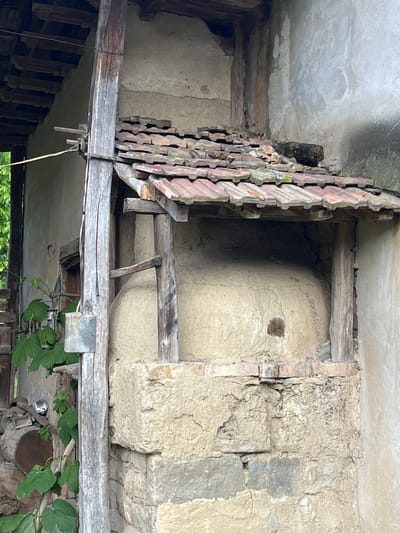 The image, originally inverted, depicts an old, rustic architectural space, likely an interior or a covered outdoor area of a rural building. The central subject is a large, rounded, light-colored structure, seemingly constructed from mud, clay, or a similar earthen material. It features a rough, textured surface with a noticeable dark brown patch on its side. This structure is integrated into a wall made of stone and mortar, with some sections of brick visible.
The space is framed by weathered wooden beams and planks, suggesting an aged but robust construction. To the left, a distinctive vertical element, possibly a decorative screen or vent, is made of numerous terracotta-colored, carved, and interlocking pieces, resembling old roof tiles or carved wood. Above, the ceiling, likely concrete or plaster, shows signs of age and wear.
In the lower right portion of the correctly oriented image, green vine foliage with broad leaves grows into the scene, partially obscuring a reflective, silvery object and a piece of cut wood or a small log. No people are present, and no specific activity is taking place. The overall impression is one of age and disuse, characteristic of traditional rural architecture in a location such as Byala Reka, Bulgaria. The lighting suggests ambient daylight. No visible text is present.