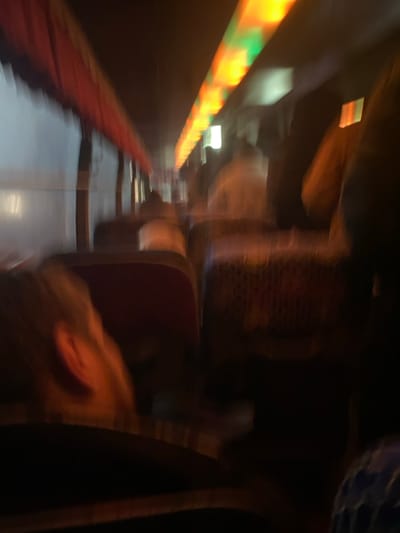 This image depicts the interior of a public transport vehicle, likely a bus or trolleybus, in motion within the city of Oktyabrskiy, Russia, under low light conditions. The perspective is from a seated position, looking forward down an aisle.

**Environment:** The vehicle's interior is dark with significant motion blur, indicating movement. Overhead, a continuous strip of illuminated panels emits orange/yellow and green light, creating a distinct ceiling glow. A red or maroon fabric-covered railing or trim runs along the upper left side of the interior. Windows on the left side of the vehicle show indistinct exterior light.

**Objects:** Rows of seating are visible. The seatbacks are upholstered in a dark, possibly maroon or red, fabric. The back of a seat in the mid-ground features a patterned or mesh-like covering.

**People:** Multiple blurred human figures are discernible, occupying seats within the vehicle. Their specific features are indistinguishable due to the blur and lack of light. In the immediate foreground, to the left, the side profile of a person's head, including a visible ear, is present. This individual appears to have light-colored hair and is wearing dark clothing, facing away from the camera. No discernible actions or interactions between individuals are visible.