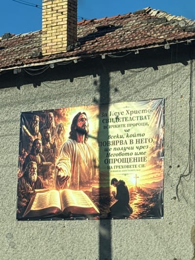 The image depicts a banner hanging on the exterior wall of a building. The banner features a religious scene with Jesus Christ and several other figures, along with text in Bulgarian. In the foreground of the banner, Jesus is depicted with a radiant halo, extending his hand. To the left, a group of men, some with beards and wearing robes, are gathered around an open book. One man appears to be writing. In the background, a landscape with a cross and a person kneeling in prayer is visible. The text on the banner translates to: "For Jesus Christ, all the prophets testify that everyone who believes in him will receive forgiveness for his sins through his name. Acts of the Apostles 10:43."
The building itself has a textured, light-colored concrete wall. A tiled roof with a brick chimney is visible at the top of the image. The overall impression is that of a religious poster displayed outdoors, likely in a village or town setting. This specific banner is located in Veselinovo, Bulgaria. The lighting suggests it is daytime, with sunlight casting shadows.