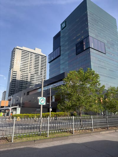 An urban street scene unfolds on a clear, sunny day in what appears to be Longueuil, Canada, featuring prominent modern architecture and street infrastructure. The photograph is taken from a slightly elevated perspective, looking across a street towards several high-rise buildings.

The dominant structure is a sleek, modern skyscraper with a facade composed primarily of dark blue-green glass panels, located in the upper left and central-left of the frame. Sections of the glass reflect the bright blue sky, while others appear darker. A distinct feature is a large, dark-framed, rectangular window or architectural opening set into a darker, non-glass portion of the building. Near the bottom-left, a white logo depicting stylized intertwined letters, likely 'P' and 'R', is affixed to a dark panel.

To the right and slightly behind this glass tower, another high-rise building with a lighter, possibly grey or off-white, facade is visible. This building exhibits a more traditional design with numerous uniform, recessed rectangular windows and a textured, stepped profile.

In the mid-right of the image, a vibrant green tree with dense foliage partially obscures the glass skyscraper. Running horizontally across the mid-right is a long, grey metal fence with a distinctive wavy, patterned design, separating a strip of greenery from an asphalt road. The bright sunlight casts crisp shadows, indicating the time of day is likely late morning or early afternoon.

Attached to a grey street light pole near the fence are two street signs. The upper sign is a circular green disc with a white arrow pointing left. Below it, a smaller green circular sign with a white 'P' signifies parking, with faint green text beneath it that potentially reads "VISITEURS". No human activity or specific events are depicted in this static urban landscape.