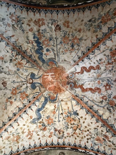 This image displays a richly decorated vaulted ceiling, likely a fresco, found within a historic building such as a church or chapel in Tepotzotlán, Mexico. The entire arched surface is covered in intricate painted motifs.
At the very center of the ceiling, a prominent reddish-orange oval or shield-like emblem draws the eye. Within this emblem, the stylized Christogram "IHS" (Iesus Hominum Salvator – Jesus, Savior of Men) is clearly visible, surmounted by a small cross. This central religious symbol is surrounded by swirling blue and reddish-brown decorative ribbons and abstract patterns. Thin, dark reddish-brown diagonal bands radiate outwards from the center, delineating sections of the vault and creating a structural framing effect.
The bulk of the ceiling is adorned with abundant stylized floral patterns and foliage. These flowers are rendered in muted tones of orange, blue, and gray, intertwined with green-gray leaves, forming a dense and organic tapestry across the light, aged cream background. Small, repeated decorative patterns, resembling scallops or stylized leaves, border some of the structural lines and sections.
Directly below the central IHS motif, a small, pale face, characteristic of a cherub or putto, is subtly depicted, appearing to gaze downwards from the celestial composition. The frescoes exhibit a decorative style reminiscent of colonial Mexican art, likely influenced by Baroque or Rococo aesthetics, yet with a distinct folk-art quality in its execution. The paint appears somewhat faded and aged, showing the texture of the underlying plaster, which contributes to its historical authenticity. No activity is taking place; the image is a static portrayal of architectural and religious artwork.