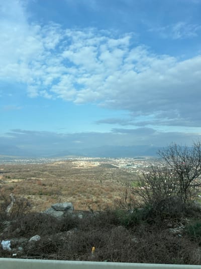 This image presents a panoramic landscape view from an elevated position overlooking the city of Gornji Kokoti, Montenegro. The immediate foreground features a white horizontal barrier, likely a guardrail, spanning the bottom of the frame. Above this barrier, the terrain is covered with dense, dry, dormant shrubbery and scattered detritus, including two prominent grey rocks centrally located and a leafless deciduous tree with dark branches on the right. Visible among the sparse vegetation are small pieces of white plastic and a yellow cylindrical object.

The midground consists of an extensive, undulating landscape dominated by numerous bare deciduous trees and shrubs, displaying a muted palette of browns and tans, indicative of winter or late autumn. A faint winding road or path is visible descending through this terrain on the far left.

In the background, the city of Gornji Kokoti sprawls across a broad plain, characterized by a concentration of urban buildings and structures. Further in the distance, mountains, some with visible snow patches on their peaks, define the horizon line, particularly towards the right side of the frame. Atmospheric haze partially obscures the most distant geographical features.

The sky occupies the upper portion of the image, displaying a blend of clear blue and various cloud formations. White cumulus clouds are scattered across the sky, with a more substantial band of grey clouds extending horizontally across the middle-right. The scene is illuminated by natural daylight. No individuals, specific actions, or interactions are discernible.