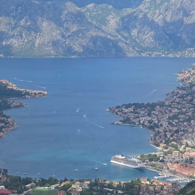 An aerial view of the Bay of Kotor, Montenegro, includes mountains to the left and developed areas on the right. The bay is filled with blue water, with several boats visible. A large cruise ship is docked near a town with many buildings and trees. Another town, also densely populated, is situated on a peninsula in the lower part of the image. The mountains are rugged and sparsely vegetated. The developed areas feature buildings with red tile roofs.