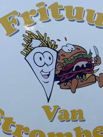 The image displays a cartoon graphic featuring a French fry character and a hamburger character running. The French fry character is white with black outlines and has large eyes and a smiling mouth, with French fries sticking out of its top. The hamburger character is brown with a bun, lettuce, tomato, cheese, and a dripping sauce, and it is holding a knife in its right hand. Both characters are depicted with legs and appear to be in motion. Above them, large yellow text with a grey shadow reads "Frituu", suggesting a name for a fast-food establishment. Below, smaller yellow text with a grey shadow reads "Van", and further below, partial yellow text with a grey shadow reads "trome be". This suggests the name of the establishment is likely "Frituu Van Trombe". The background is a plain white surface. The context provided indicates this image is located in Strombeek-Bever, Belgium, likely representing a local business or advertisement.