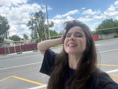 A young woman with long, dark, wavy hair is captured in a selfie. She is wearing a dark blue t-shirt and has her right hand behind her head. Her eyes are closed and she is smiling broadly, showing her teeth. She is wearing subtle makeup, including eyeshadow. The background indicates an outdoor urban environment with a road, sidewalk, a fence, some houses, and trees. The sky above is blue with scattered white clouds. A yellow pedestrian crossing sign is visible to the left of the woman.