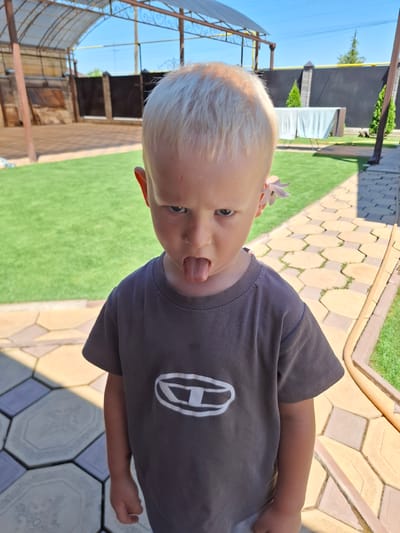 In the city of Kara-Balta, Kyrgyzstan, on a sunny day, a young Caucasian boy with blonde hair and blue eyes stands on a patterned stone walkway. He is wearing a dark gray t-shirt with a white logo. The boy is sticking his tongue out with a mischievous expression on his face, and a light purple flower is tucked behind his right ear. The background features a green lawn, a covered patio area with a transparent roof, and a fence. The lighting suggests it is daytime, with shadows cast on the walkway.