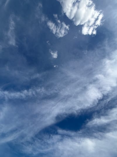 The image captures a wide-angle view of a deep blue sky filled with a variety of clouds. On the left side of the frame, a cluster of bright white, cumulus clouds stands out against the darker blue. These clouds have well-defined shapes and appear to be illuminated by direct sunlight.
Further into the sky, a dense formation of wispy, feathery cirrus clouds stretches diagonally across the frame from the top right to the bottom left. These clouds are translucent and appear thinner, suggesting they are at a higher altitude. The way they are shaped and spread creates a sense of movement and depth. In between these formations, patches of lighter blue sky are visible.
The lighting suggests it is daytime, likely mid-day given the brightness of the cumulus clouds. The scene depicts a natural outdoor setting, specifically looking up at the sky. There are no people, objects, or text visible in the image. The overall impression is one of a clear, but cloudy, day. The location context provided is Мартемьяново, Russia, which adds a geographical setting to the observation of the sky.