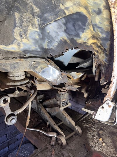 The image depicts the underside of a vehicle that has undergone significant damage, primarily due to rust and structural deterioration. A hydraulic car jack is visible, supporting the vehicle, indicating it is either undergoing repair or has been immobilized. The chassis is extensively corroded, with large holes revealing the underlying structure. Various metal components, some appearing to be part of the suspension system, are visible. The ground beneath the vehicle is covered in dirt and debris. The scene appears to be an indoor or sheltered location, with a brick wall visible in the background. There are no people present in the image. The context of Maladzyechna, Belarus is not visually evident from the provided image.