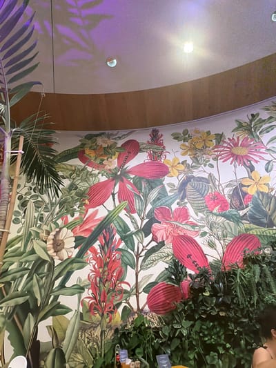 The image depicts an indoor environment with a curved architectural design. The ceiling is light-colored and features multiple recessed circular light fixtures; one emits bright white light and glare, while another reflects a purplish-blue hue, casting shadows of plant foliage onto the surface. Below the ceiling, a curved wooden panel functions as a bulkhead or trim. Below this panel, a large-scale botanical mural covers the wall, depicting a dense arrangement of tropical flora. The mural features prominent red, pink, and yellow flowers, including hibiscus-like forms and spiky inflorescences, interspersed with various green leaves on a light background. In the foreground, particularly along the bottom and right side, live or artificial greenery, including ferns and broadleaf plants, is densely arranged, obscuring the lower portion of the mural. On the far left, a tall potted plant with palm-like fronds and bamboo-like support stakes is visible. In the lower right corner, the upper right shoulder, arm, and a portion of the head of an individual with dark hair, wearing a dark, thin-strapped garment, are partially visible. Indistinct objects are present on a surface, possibly a counter or table, in the lower left foreground. The image does not provide information to identify the specific location within Săftica, Romania.