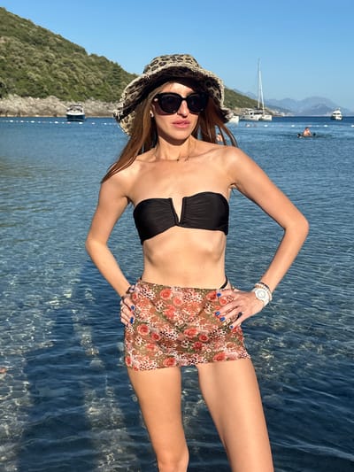The image depicts a sunlit coastal scene in Kubasi, Montenegro. An adult female stands in the foreground, facing the camera, with clear, shallow, turquoise water reaching her upper thighs. She wears a strapless black bikini top, a sheer high-waisted mini-skirt featuring a floral and leopard print pattern in warm tones, and a distressed leopard print bucket hat. Dark-lensed sunglasses obscure her eyes. A delicate gold-colored necklace with a small pendant is visible on her chest. Her left wrist displays a white wristwatch along with several white beaded and metallic bracelets. Her fingernails are painted a vibrant blue, and she wears two rings on her right hand, which is placed on her hip. Her lips are slightly parted.
In the midground and background, the calm bay features several boats: a larger white motor yacht is positioned to the left against a green mountain, a white sailboat with a tall mast is visible towards the center-right, and additional smaller motorboats are scattered further in the distance. Several indistinct human figures are seen swimming or wading in the water in the midground, behind a visible line of white buoys. The bay is framed by steep, lush green, tree-covered mountains, with more distant, hazy blue mountains under a clear, bright blue sky. The scene is brightly illuminated by natural sunlight.