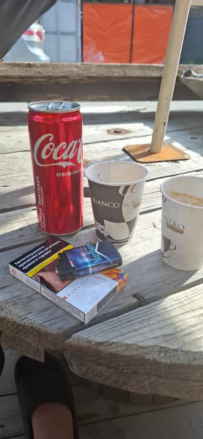 A Coca-Cola can, two paper cups, and two packs of cigarettes are arranged on a wooden table in an outdoor setting. The Coca-Cola can is red with white lettering. One cup is black and white with "BIANCO & NERO" printed on it. The other cup is white with faint brown markings and "NERO" visible. A Marlboro cigarette pack with blue and silver accents is placed on top of a Karelia cigarette pack, which has orange and white packaging. In the background, an orange tarp covers a structure, and a portion of a car is visible. The overall scene suggests a break or a casual gathering.