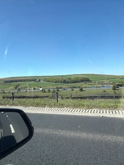The image, when viewed in a landscape orientation, presents a bright, sunny daytime scene of a rural or semi-rural landscape, likely captured from a moving vehicle. A clear, vibrant blue sky occupies the top portion of the frame, showing faint white streaks, possibly contrails or lens flares.
In the immediate foreground, towards the right side, a dark asphalt road surface is visible, bordered by a concrete drainage channel and a narrow strip of green grass. Beyond this, the mid-ground reveals a wide, verdant valley. A multi-lane road or motorway cuts through this valley, with several vehicles, including a white van, discernible on it. The road is flanked by patches of green trees and extensive fields, which stretch towards distant, undulating green hills. Scattered buildings, possibly farmhouses, are dotted across the landscape in the distance. A body of water, resembling a pond or small reservoir, is also visible within the valley.
The lower right corner of the image prominently features the black casing and reflective surface of a car's side mirror. The mirror reflects a section of the road directly behind the vehicle, including clear white lane markings. A small, white rectangular sticker or label is adhered to the mirror, but any text on it is illegible. The scene evokes a sense of travel through the scenic, open countryside, consistent with the rolling hills and green expanses found in areas like Calderdale, United Kingdom.