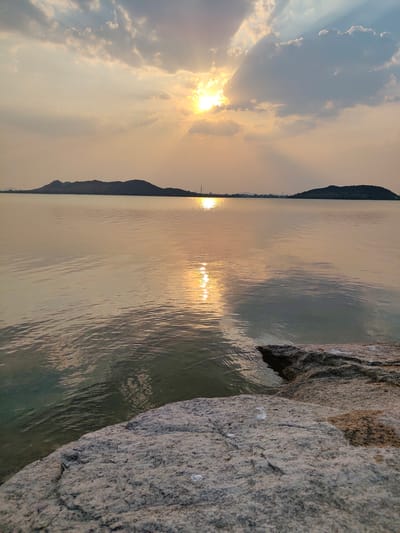 The image depicts a serene lake scene in Kammavaripalli, India, likely at sunset. The sun is partially obscured by clouds, casting a warm, golden light across the water and creating sunbeams. The water is relatively still, with gentle ripples reflecting the sunlight. In the distance, two hills or landmasses are visible, silhouetted against the horizon. A few structures, including what appears to be a tower, are visible on the left side, slightly further back. In the foreground, a large, textured rock formation juts out into the lake. The sky is filled with clouds, ranging from light, wispy formations to darker, more substantial ones. The overall color palette is dominated by shades of gold, gray, and blue. The scene is peaceful and quiet, with no people visible.