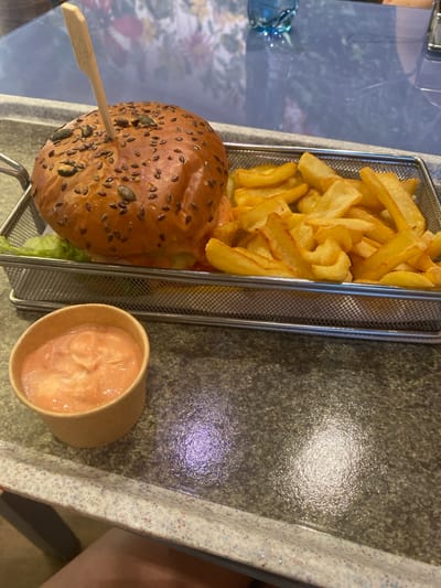 The scene, located in Săftica, Romania, depicts a meal served on a dark, speckled tabletop. A burger, held by a vertical wooden skewer, rests within a rectangular metal mesh basket. The burger features a brioche bun topped with sesame, flax, and pumpkin seeds. Visible components inside the bun include green lettuce and a reddish sauce. Alongside the burger in the same basket is a portion of golden, thick-cut french fries. To the lower left of the basket, a small, light brown paper cup contains a light orange-pink dipping sauce. The tabletop exhibits areas of wetness and reflections of overhead lighting. In the blurred background, a blue glass bottle and a floral-patterned surface are visible. A human knee is partially visible at the bottom left of the frame.