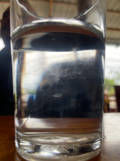 A close-up, slightly upward-angled shot shows a clear glass filled with water sitting on a wooden surface. The glass is mostly in focus, revealing some streaks and imperfections on its surface, as well as ripples and bubbles at the bottom. The water level is about two-thirds of the way up the glass. Behind the glass, a dark, blurred shape is visible, possibly a person or an object. The background is out of focus, showing hints of an outdoor setting with greenery and what appears to be a thatched roof, suggesting a rustic or natural environment. The lighting is somewhat diffused, creating reflections and refractions within the glass and water. No specific landmarks or indications of Julikeude Dua, Indonesia, are present in the image.
