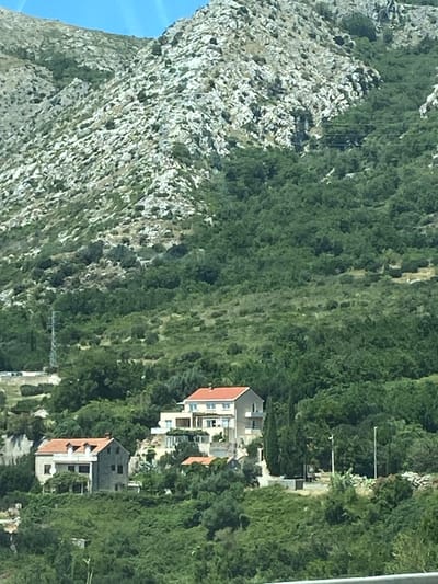 The image shows a hillside with several houses nestled amongst lush green vegetation. In the foreground, a cluster of houses is visible, one with a red tile roof and dormer windows, another larger house with a light-colored facade and a similar red tile roof, and a smaller house with a red tile roof to the right. The houses appear to be located in a somewhat remote or rural setting. The background is dominated by a steep, rocky mountain slope covered with sparse greenery and scrub. A tall, thin electrical pylon is visible on the left side of the image, and two tall, slender poles with what appear to be lights or signs are located further into the landscape on the right. The sky is a bright, clear blue, suggesting a sunny day. The image captures a scene in the vicinity of Plat, Croatia, a coastal village.