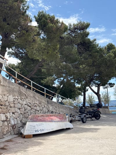 The image shows a scene on a coastal path in Dugi Rat, Croatia. The path is lined with a stone wall on the left, with a metal railing in front of it. The wall is overgrown with vegetation and has graffiti on it. There are two large pine trees along the path, with their branches stretching over the wall. A small boat is lying overturned on the path, and a black motorcycle is parked next to it. The motorcycle has a trailer attached to it, and both are parked on the paved path. The path is deserted, and the sky is clear and blue. The image is taken from a low angle, looking up towards the trees. The scene is captured in daylight.