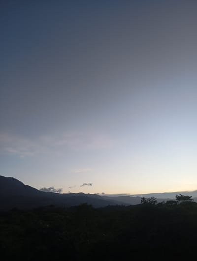 The image depicts a landscape in Bukitmeusara, Indonesia, taken during twilight or dawn. The sky transitions from a deep blue at the top to a lighter blue/white near the horizon. A range of mountains is silhouetted against the sky. A dense forest or vegetation occupies the foreground, appearing dark and undefined due to the low light. There are clouds visible at the horizon. No people are visible.