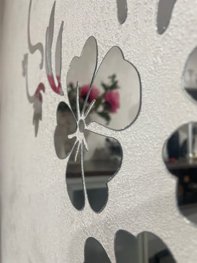 The image is a close-up, shallow depth-of-field shot of decorative mirrored flower stickers adhered to a textured, light-colored wall. The stickers are silver and have a reflective surface, showing distorted reflections of the surroundings. In the center, a large mirrored flower is prominent, with its petals and stamen clearly visible. A reflection of pink flowers and green leaves is visible within one of the petals. Other mirrored flower shapes are visible partially in the frame, some in focus and some blurred due to the shallow depth of field. The wall itself has a granular texture, possibly wallpaper or painted plaster. The lighting suggests an indoor environment, possibly a domestic setting. The location information "Aropakkuzi, Russia" is not visually supported by the image content.