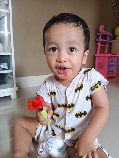 A young child, appearing to be a toddler, is seated on the floor, holding a piece of watermelon on a green utensil. The child is wearing a white onesie with a pattern of black and yellow Batman logos. The child's face shows some red residue around the mouth, consistent with eating the watermelon. In the background, to the left, is a white cabinet with glass doors. To the right, a child-sized pink and purple play kitchen is visible. The flooring appears to be tile. The overall scene suggests a casual indoor setting, likely a home environment. While the image itself does not contain any explicit geographical markers, the request mentions the location of Cibungbulang, Indonesia.