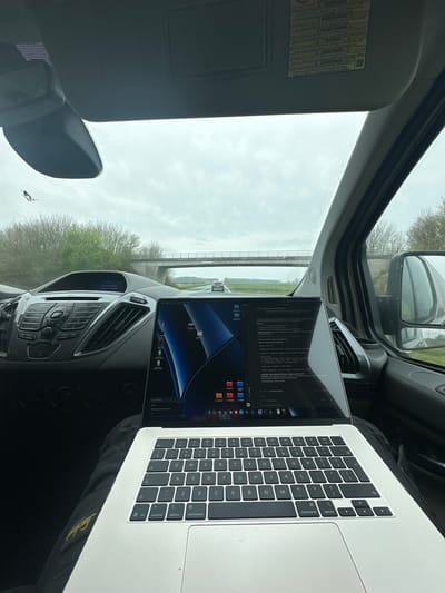 The image depicts an interior view from a vehicle, looking forward through the windshield towards a road and landscape in Druten, Netherlands. In the immediate foreground, a silver-colored MacBook laptop rests on a person's dark pants or lap. The laptop screen is illuminated, displaying a macOS desktop with a blue and black abstract wallpaper, a dock populated with numerous application icons, and two open windows. One window appears to be a document or text editor with content predominantly in Cyrillic script, while the other window's content is less discernible but appears to be a file browser or web page. An "iCloud" notification is visible near the bottom left of the screen.

The vehicle's dashboard is visible behind the laptop, featuring a dark gray plastic construction. The central console includes an audio unit with a small monochrome display indicating "07 audio," "Play music from," and "Bluetooth device." Below this are physical buttons and knobs for audio control, and to the right, vertical air vents. A rearview mirror hangs from the ceiling, and the passenger-side sun visor is visible above it, bearing a multi-language warning sticker, including the Dutch word "WAARSCHUWING" (warning). A small crack or defect is present on the upper left of the windshield. The passenger-side door panel and a portion of the side-view mirror, reflecting the vehicle's side, are visible on the right.

The exterior environment, observed through the windshield, shows a two-lane road stretching into the distance under an overcast sky. Green trees and brush line the sides of the road. A multi-lane concrete bridge spans the road in the mid-distance. Another vehicle is visible in the distance on the road, traveling in the same direction. No individuals are directly visible, though a person is implied to be operating the laptop and holding the camera within the vehicle.