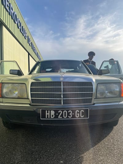The image captures a light green Mercedes-Benz sedan, model W126 S-Class, with French license plate "HB-203-GC," parked on dark asphalt. The vehicle's driver-side and front passenger-side doors are open. A person is seated inside the driver's compartment, partially visible. Standing centrally behind the car's hood, a male individual with dark, curly hair, dressed in a suit jacket and tie, leans into the vehicle between the open doors. To the right, partially obscured by the open passenger door, another individual is seen holding a camera, facing towards the car. In the background, under a bright blue sky with scattered clouds, is a large, light green hangar building displaying the text "AEROCLUB DE VIENNE." This location is identified as the Aerodrome de Vienne - Chonas (LFHH) in Chonas-l'Amballan, France.