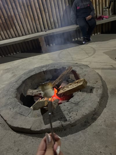 An image taken at night depicts a stone fire pit with burning wood. A person holds a skewer with food over the flames. Another person sits nearby. The location is Pacarán, Peru. The environment includes the structure with bamboo roof and concrete walls.