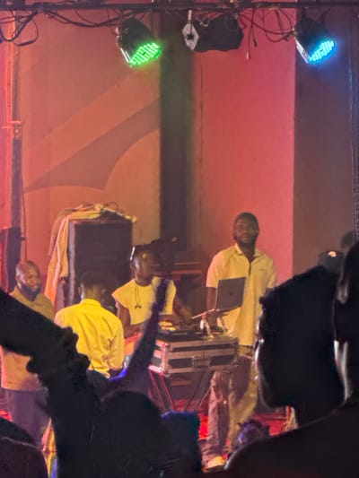The image captures an indoor event, likely a performance or concert, illuminated by stage lighting in Ife, Nigeria. The scene is dark, suggesting an evening or night event.

Several individuals are visible, primarily operating stage equipment. In the central area, a man in a light-colored shirt is positioned behind what appears to be a mixing console or audio equipment, looking towards a laptop placed in front of him. Further back and to the left (when viewed upright), another person in a light shirt is partially visible, also interacting with equipment. In the lower right foreground, a person wearing a yellow shirt is seen. Dark, out-of-focus shapes in the immediate foreground suggest the viewpoint is from within an audience or a side-stage area.

Prominent stage lights add color to the scene. A blue LED light is mounted on a stand in the upper left, while a green LED light is visible on another stand in the middle left. Numerous cables connect the lighting and other equipment. The background consists of a reddish-orange wall or screen, likely a stage backdrop, with faint, subtle geometric patterns. No discernible text is visible in the image, and due to the distance and lighting, specific emotions of the individuals are not apparent. The overall atmosphere is focused and operational, centered around the event setup.