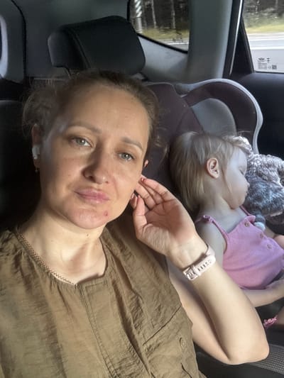 A woman and a sleeping child are in the back seat of a car. The woman is in the foreground, looking at the camera. She has light brown hair pulled back and is wearing a brown top and a pink watch. She has earrings and an earbud in her left ear. A reddish mark is visible on her chin. The child, a toddler, is in a car seat behind the woman and is wearing a pink top. A stuffed animal is next to the child. The car interior is visible, including a car seat and headrests. Through the window, trees and a road are visible, suggesting a rural or suburban setting. The lighting is somewhat dim, indicating it could be daytime with overcast skies or late afternoon. There is no specific information visible in the image that would indicate the location as Yus'ki, Russia.