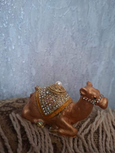 A close-up, eye-level shot shows a decorative camel figurine with a bronze finish and ornate embellishments. The camel is adorned with a jeweled halter and a saddle blanket encrusted with small, clear and pink rhinestones, along with a single pearl accent on the blanket. Its posture suggests it is standing. The figurine is positioned on the right side of the frame, leaning against a textured cylindrical object wrapped in thick, beige rope, which creates a rustic and tactile element. To the left of the camel, the background is a softly lit, textured surface with a light blue-grey hue. This surface appears somewhat uneven, with subtle variations in tone and texture, possibly suggesting a stone or textured fabric. The lighting is soft and diffused, with no harsh shadows, creating a calm and contemplative mood. The image is static, with no discernible activity or event taking place, focusing solely on the aesthetic details of the decorative object and its immediate surroundings. There is no visible text in the image. The overall impression is one of intricate craftsmanship and a gentle, serene atmosphere.