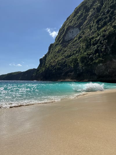 The image shows a beach scene in Bunga Mekar, Indonesia. A cliff covered in dense green vegetation dominates the left side of the image. The right side displays a sandy beach meeting the turquoise water of the ocean. A small wave is breaking on the shore. The sky is visible in the upper left corner with a few small clouds. The image is rotated 90 degrees clockwise.