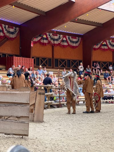 The image depicts an outdoor arena, likely within the city of Collendoorn, Netherlands, where a performance or reenactment is taking place. The scene is set in what appears to be a themed venue, possibly a Wild West attraction, judging by the attire of the main subjects.

In the foreground, three individuals are dressed in Native American-inspired costumes. The person on the left wears a brown buckskin outfit and a headdress adorned with fur and feathers. They are holding a large feathered shield-like object. The middle individual is also in brown buckskin, with a red headband and face paint. They appear to be gesturing towards the person on the right, who is wearing a similar buckskin outfit with intricate beadwork and a feathered ornament. The ground of the arena is covered in sand or fine gravel.

Behind the performers, wooden barricades and fencing are visible, separating the performance area from the audience. A crowd of spectators is seated on tiered wooden benches in the background. They are of various ages and are casually dressed. Some are looking towards the performance, while others are engaged in conversation.

The backdrop of the arena is a wooden structure with large, dark-stained beams and plywood walls. American-style bunting, featuring red, white, and blue stripes with stars, is draped along the upper edges of the structure, suggesting a patriotic or thematic element to the venue. Strings of colored lights, some red and some yellow, are strung across the ceiling beams. The overall atmosphere appears to be that of a family-friendly entertainment event.