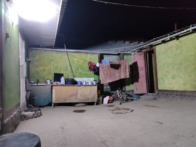 The image shows an outdoor kitchen area at night in Ismoili Somoni, Tajikistan. A bright overhead light illuminates the scene, revealing a rough, concrete floor with several damp patches. To the left, a section of a green wall has a white door. Adjacent to this is a countertop area with a sink, various pots, kettles, and a large white jug. In the center, a rustic wooden counter or cabinet stands, with a spare tire visible underneath. Hanging on a clothesline above are several pieces of clothing, including a red and blue shirt and a striped cloth. To the right, a bicycle is propped up near a red wooden door. The walls are painted a mottled green, with a stone base running along the bottom. The overall impression is one of a functional, somewhat cluttered outdoor living space.