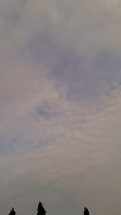The image shows a sky with light, muted colors and a mix of clouds. Three tree tops are visible in the bottom right corner. Based on the location provided, it is a daytime view of the sky and trees in Umuigbekwo, Nigeria.