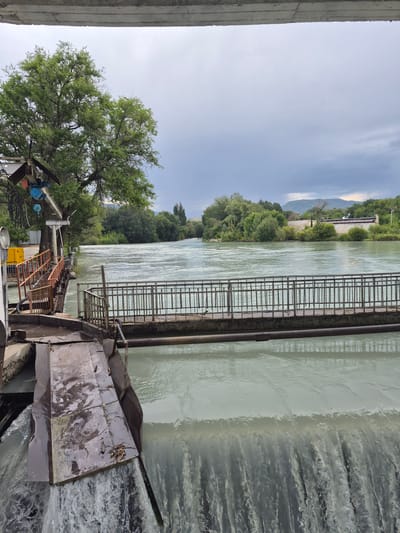 The image shows a wide, calm river flowing past a concrete structure with a metal railing. On the left side of the river, there is a building with a yellow generator and orange railings. A large green tree stands behind the building. The river continues into the distance, bordered by lush green trees and vegetation. In the background, mountains are visible under a cloudy sky. The overall scene appears to be a scenic view of a river and its surroundings in a natural setting, potentially in or near the city of Tekeli, Kazakhstan, given the context.