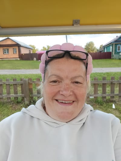 This is a close-up, selfie-style photograph of a woman outdoors. She is wearing a light-colored hooded sweatshirt and a pink, pom-pom-style headband. Her black-rimmed glasses are perched on top of her head. The woman is smiling broadly, her eyes crinkled at the corners, suggesting a happy or amused expression. She has fair skin with freckles and her hair is light blonde.

The background of the image provides context for the setting. Behind the woman, there is a wooden fence and then a grassy area. Further back, two houses are visible, one with a light brown exterior and a darker brown roof, and another with a blue and green facade. The houses appear to be in a rural or village setting. The sky above is overcast, suggesting it might be a cloudy day, possibly after rain, as there appear to be some water droplets on a surface above the woman's head. The overall lighting suggests it is daytime, likely afternoon given the soft light. The presence of the houses and the fence in a grassy area indicate a residential location in a village, consistent with the provided location context of Smolino, Russia. No visible text is present in the image.