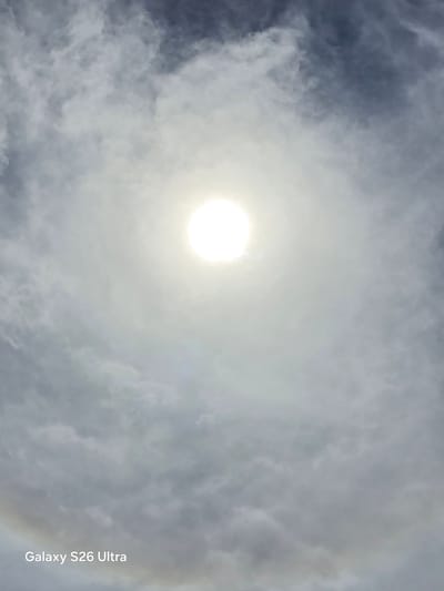 The image shows a cloudy sky with the sun prominently visible in the center, creating a halo effect around it. The sun appears as a bright white disc, and a faint rainbow or halo ring is visible in the lower left corner. The clouds are varied in texture and color, ranging from light gray to darker blue-gray. The bottom left of the image contains a watermark that reads "Galaxy S26 Ultra". No people or specific landmarks are visible to indicate a precise location within West Hartford, United States of America. The scene depicts a natural atmospheric phenomenon.