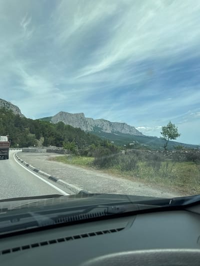 The image shows a view from inside a vehicle, looking out at a landscape with a mountain range, a road, and the sky. The sky is light blue with streaky white clouds. A mountain range stretches across the middle of the frame, with a rocky peak visible. Vegetation covers the lower slopes of the mountains. A paved road runs parallel to the mountain, with a white and black painted barrier along the edge. In the foreground, the interior of the vehicle is visible. The photo was taken in Форосский поселковый совет, Ukraine.