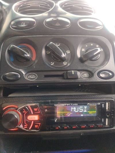 The image presents a detailed view of a vehicle's dashboard interior in the city of Ortiugil, Uzbekistan. The upper section consists of a black plastic climate control panel featuring four circular air vents along the top edge, partially obscured by direct light. Below these vents are three black rotary knobs: the left knob, controlling temperature, is set toward the blue (cooler) indication; the central knob, for airflow direction, is positioned to direct air primarily to the upper vents; and the right knob, for fan speed, is set to a medium setting. A series of black push-buttons are present, including one for headlights (top-left) and a hazard warning light button (top-center). A slider switch, controlling air recirculation or fresh air intake, is positioned to the right.
Below the climate controls, an aftermarket "Pioneer.GB RADIO/USB/TF MP3 PLAYER" car stereo unit is installed. The unit's black faceplate exhibits a textured, carbon fiber-like finish. On the left side, a rotary volume knob is illuminated with an orange ring, and the adjacent "MUTE" button glows red/orange, indicating active muting. The central digital display shows the text "MUSI" in white characters, accompanied by an active, multi-colored graphic equalizer visualization. The right side of the stereo unit features two USB ports and a TF (MicroSD card) slot. A row of numbered function buttons lines the bottom edge of the stereo. Fine particulate matter is visible on all dashboard and stereo surfaces. No individuals are depicted in the scene.