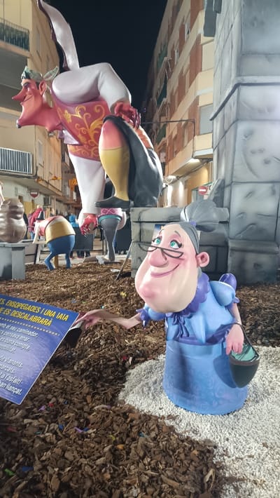 The image displays a nighttime street scene in El Port De Sagunt, Spain, featuring a large, elaborate sculpture installation typical of a Fallas celebration.
In the immediate foreground on the right, a sculpted figure depicts an elderly woman with light skin, black spectacles, and a cheerful expression. She wears a light blue dress with a frilly collar and a patterned darker blue skirt. A small dark green bucket, containing light green material, is held in her left hand. Her right arm is extended towards a partially visible blue and white informational sign located on the left side of the frame, which displays text in Valencian/Spanish, including the word "ENSOPEGADES."
The ground around these figures is covered with a layer of dark brown wood chips, interspersed with small colorful pieces of debris. The elderly woman figure stands on a distinct patch of white pebbles within the wood chips.
Dominating the upper midground is a significantly larger sculpted male figure with light skin, a prominent hooked nose, and a wide grin. He is dressed in a white shirt, white pants, a red vest with gold scrollwork, and a light green bandana. His left hand, detailed with red, is raised, while his right hand holds a dark, elongated object with yellow and pink accents, resembling a fish or similar item. His body is depicted in a bent-forward posture with legs extended.
Further in the midground, other smaller sculpted figures are visible: one figure in blue pants and a beige shirt is bent over, facing away from the viewer. Another dark brown, amorphous sculpture rests on a gray pedestal to the left.
The background consists of multi-story residential buildings with brick facades, balconies, and windows, characteristic of a city street. The scene is illuminated by artificial light sources, casting shadows and highlighting the sculptures against the dark night sky.