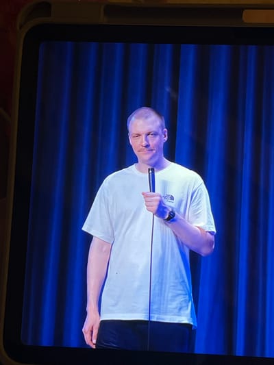 A man wearing a white t-shirt and black pants stands on a stage, holding a microphone. He has short, light-colored hair and a mustache. A blue curtain serves as the backdrop for the scene. The image is framed by a yellow border, suggesting it is a picture taken from a screen, likely a tablet or phone. There are no visible landmarks or context to place this scene in Genemuiden, Netherlands.