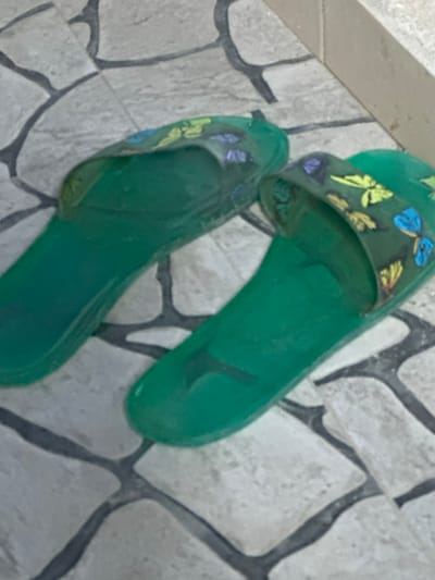 Two green, translucent jelly sandals with butterfly designs on the straps sit on a light gray tile floor with dark gray grout. The sandals appear slightly worn. The tile pattern consists of irregularly shaped stones. The upper left of the image shows a plain, lighter gray surface. The lighting is slightly dim, giving the sandals a muted color.