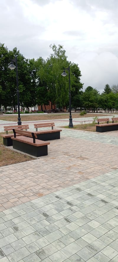 The image depicts an outdoor public area, likely a park or pedestrian zone in the city of Kirsanov, Russia. In the foreground, there are several modern wooden benches with black bases arranged on a paved pathway. The pathway is composed of light-colored rectangular tiles in a repeating geometric pattern, with sections of different colors, suggesting a designed walkway.
Behind the benches, a line of trees forms a backdrop, with a more densely wooded area further in the distance. Two ornate black lampposts with vintage-style lamps stand tall, one closer to the foreground and another further back. The sky above is overcast, with a muted, grey cloud cover.
In the middle ground, to the left, a wooden structure, possibly a pavilion or gazebo, is visible. Several more benches are situated around this structure. A few indistinct figures can be seen seated on these benches, suggesting the presence of other park visitors. The ground between the paved areas and the trees is covered in grass and dry foliage. The overall impression is one of a quiet, functional urban green space on a cloudy day.
