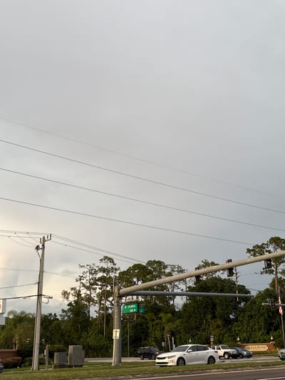 The image shows a street scene in Bradenton, Florida, United States of America. The sky is overcast, with some visible power lines stretching across the frame. In the foreground, there is a street with several cars, including a white Kia Optima, a white SUV, a dark colored SUV, and a dark colored sedan. A street sign indicates "CREEKWOOD FARM 70". Trees and foliage line the background, with a sign for "CREEKWOOD" visible. Traffic lights and utility poles are also present. The scene appears to be a typical suburban intersection on a cloudy day.